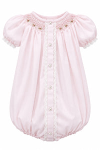 A light pink baby smocked bodysuit with short puff sleeves, lace trim, and a snap-button front, hanging on a white hanger against a neutral gray background.