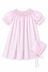 A light pink baby smocked dress with short ruffled sleeves and a matching bonnet, shown on a hanger against a neutral gray background with an inset image of the bonnet.
