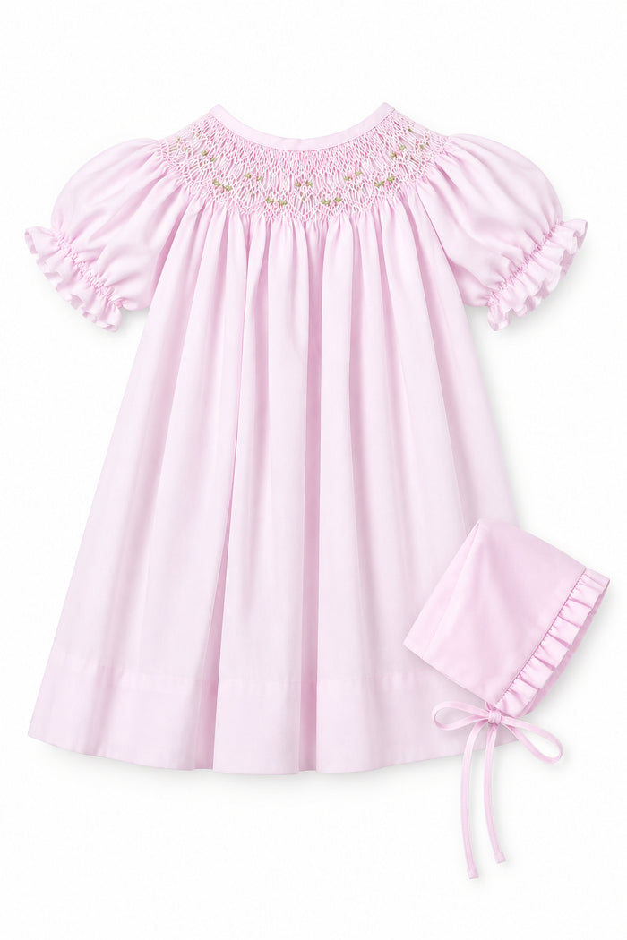 A light pink baby smocked dress with short ruffled sleeves and a matching bonnet, shown on a hanger against a neutral gray background with an inset image of the bonnet.