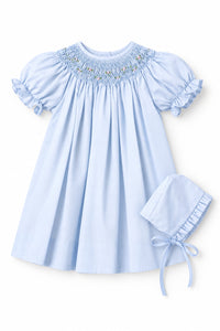 A light blue baby girl's dress with short puff sleeves and a smocked yoke, shown on a hanger with an inset image of a matching bonnet.