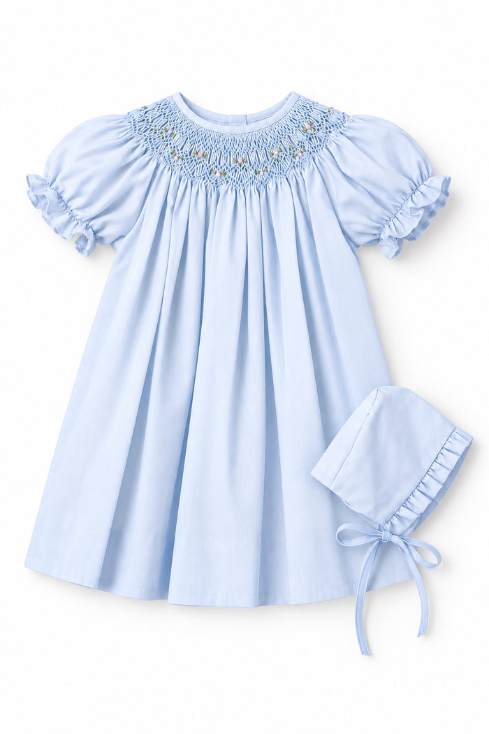 A light blue baby girl's dress with short puff sleeves and a smocked yoke, shown on a hanger with an inset image of a matching bonnet.