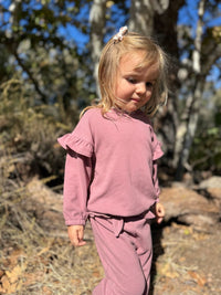 A young girl with blonde hair wearing a dusty rose two-piece loungewear set, consisting of a long-sleeve top with ruffle details and matching sweatpants, standing outdoors in a wooded setting.
