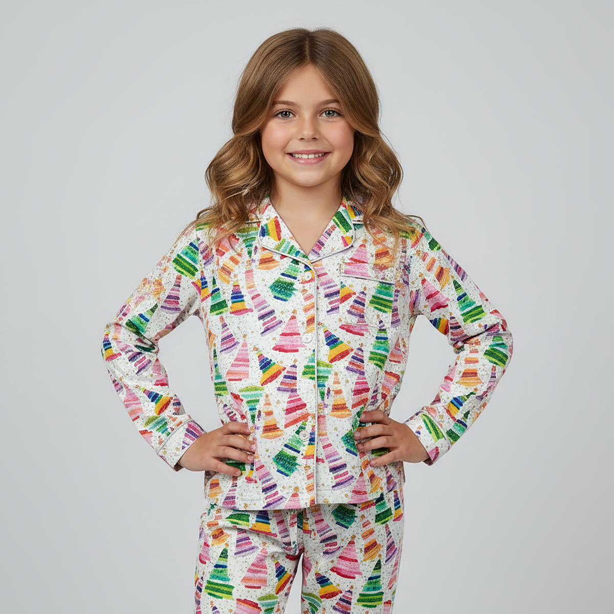 A young girl with wavy light brown hair stands against a light gray studio background wearing a long-sleeve pajama top and matching pants with a colorful crayon print on a white base.