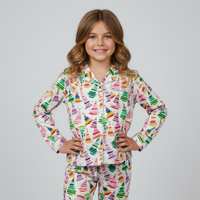 A young girl with wavy light brown hair stands against a light gray studio background wearing a long-sleeve pajama top and matching pants with a colorful crayon print on a white base.