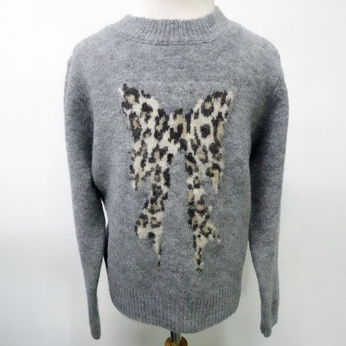 A gray knit sweater displayed on a mannequin, featuring a large leopard-print bow motif knitted across the chest.