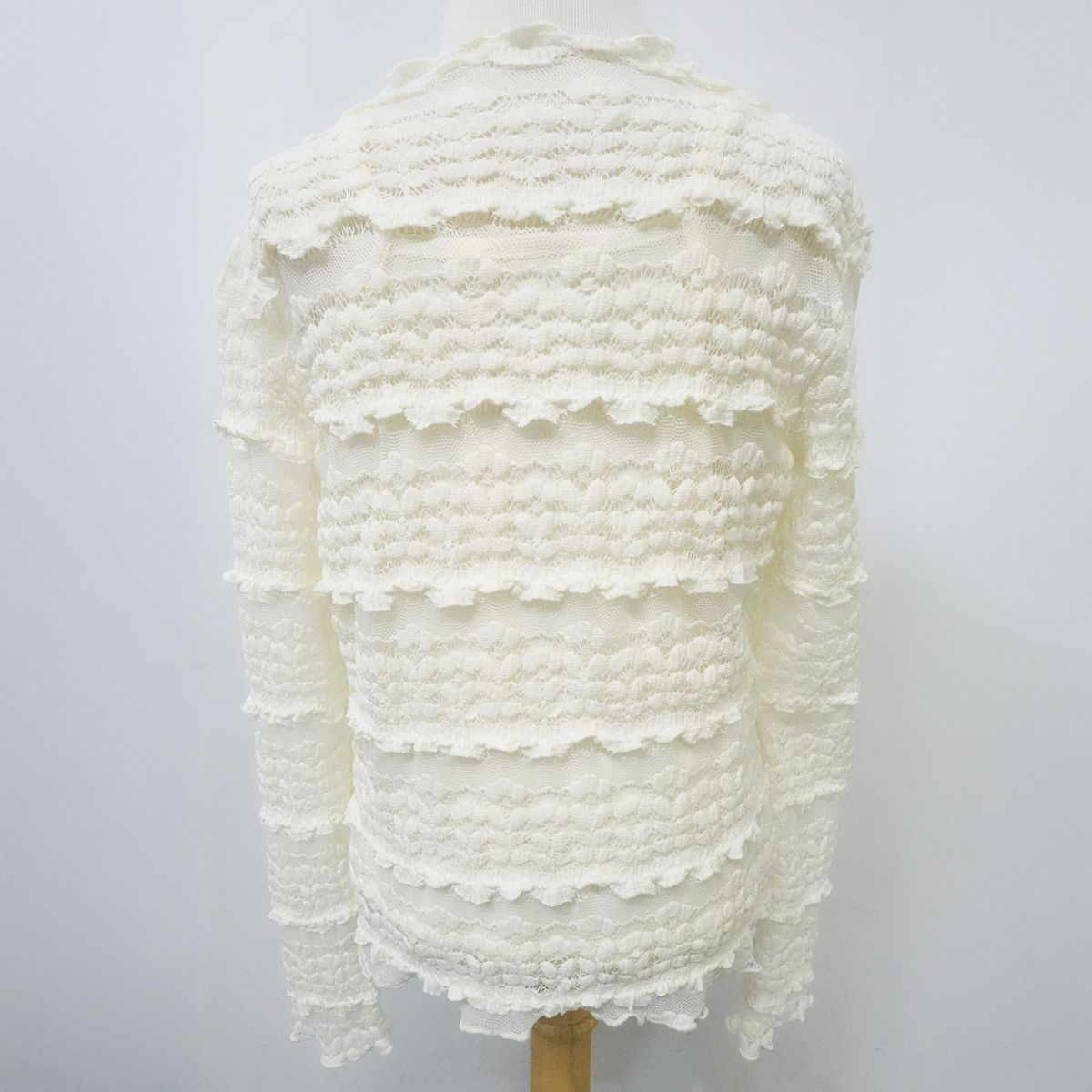 An off-white textured knit sweater with horizontal ruffled tiers, displayed on a dress form against a light gray background.