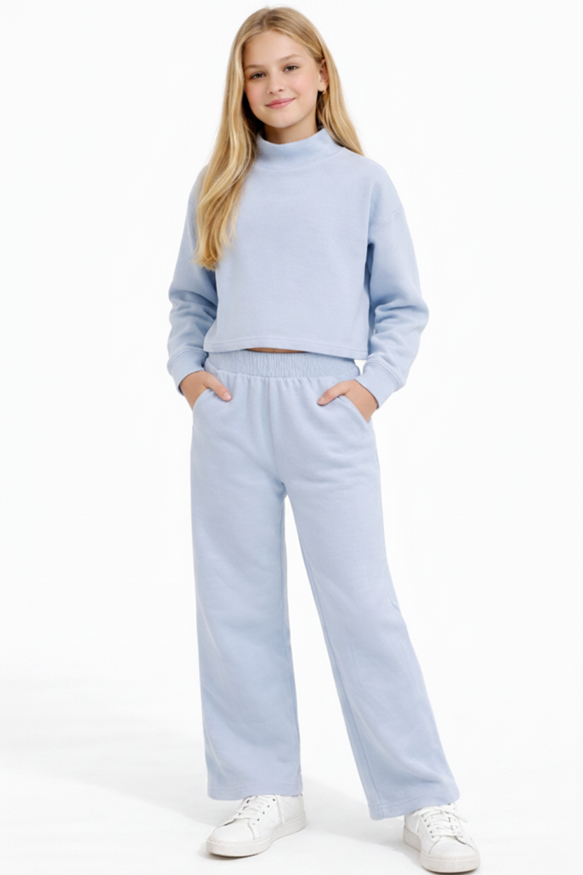 A young girl modeling a light blue two-piece lounge set consisting of a cropped mock-neck sweatshirt and matching wide-leg sweatpants, paired with white sneakers on a white studio background.