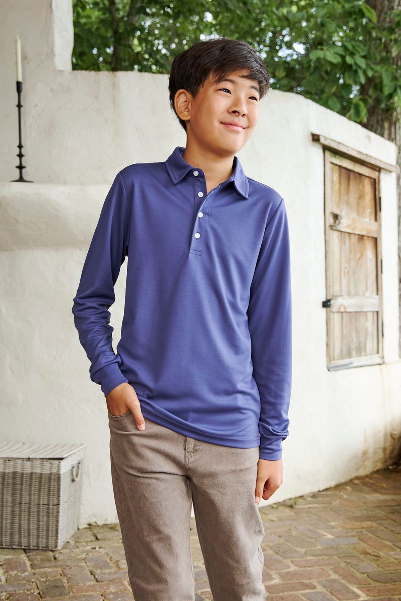 A young boy wearing a long-sleeve polo shirt in a dusty periwinkle blue color, paired with brown pants, standing outdoors against a white stucco wall.