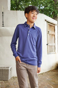 A young boy wearing a long-sleeve polo shirt in a dusty periwinkle blue color, paired with brown pants, standing outdoors against a white stucco wall.