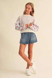 A young girl modeling a cream and lavender striped knit sweater with purple floral patterns on the sleeves, paired with a blue denim mini skirt and white sneakers, against a solid beige background.