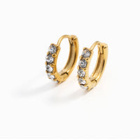 A pair of gold huggie hoop earrings with a row of clear rhinestones on the front, displayed on a clean white background.