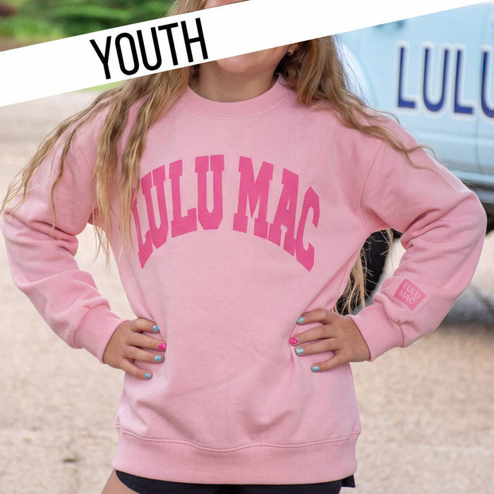 A youth model wearing a light pink crewneck sweatshirt with 'LULU MAC' printed in dark pink arched letters across the chest. The photo is taken outdoors on a sandy surface with a blue vehicle in the background, featuring a 'YOUTH' banner overlay in the top left.