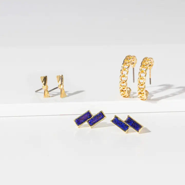 A set of gold-toned statement earrings displayed on a clean white surface, including textured gold hoop earrings, gold wedge stud earrings, and gold rectangular stud earrings with deep blue inlay.