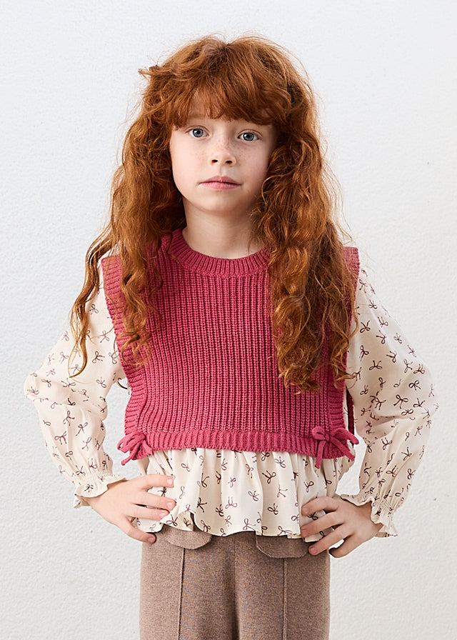 Young girl wearing a pink knitted top over a white blouse with floral details on a light gray background