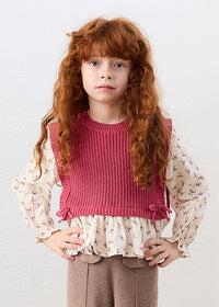 Young girl wearing a pink knitted top over a white blouse with floral details on a light gray background
