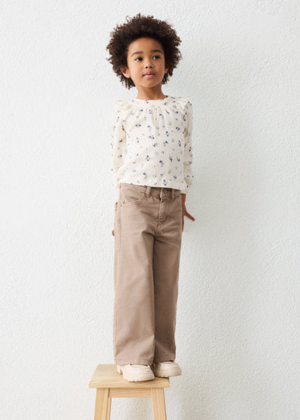 A young child with curly dark hair stands on a small wooden stool against a white textured wall, wearing a cream long-sleeve blouse with a small floral print and beige wide-leg pants.