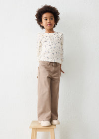 A young child with curly dark hair stands on a small wooden stool against a white textured wall, wearing a cream long-sleeve blouse with a small floral print and beige wide-leg pants.
