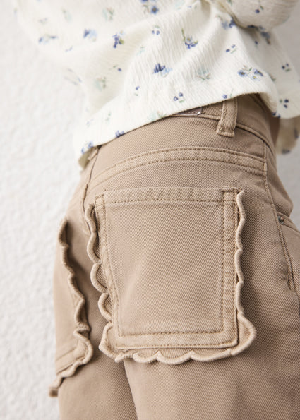 Close-up of a child wearing beige pants with scalloped edges around the back pockets, paired with a cream floral top against a light textured wall.
