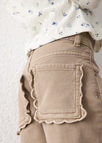 Close-up of a child wearing beige pants with scalloped edges around the back pockets, paired with a cream floral top against a light textured wall.