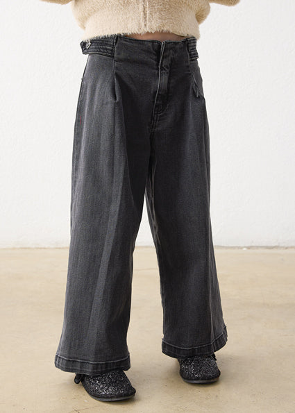 A person wearing dark charcoal gray wide-leg jeans with pleats, paired with a cream fuzzy cropped sweater and black glittery shoes, standing against a plain off-white wall and light beige floor.
