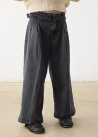 A person wearing dark charcoal gray wide-leg jeans with pleats, paired with a cream fuzzy cropped sweater and black glittery shoes, standing against a plain off-white wall and light beige floor.