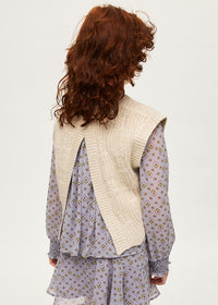 Person wearing a beige knitted vest over a patterned dress on a plain background