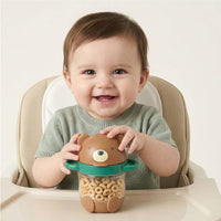 A baby sitting in a high chair holds a brown bear-shaped sippy cup filled with cereal. The cup has a green rim and handle, and the baby wears a light green knit top against a plain light background.