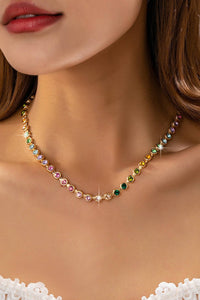A close-up of a model wearing a delicate gold necklace adorned with small, round, multicolored gemstones. The necklace is styled on a model's neck, with her lips, neck, and upper chest visible, wearing a white lace bralette at the bottom edge.