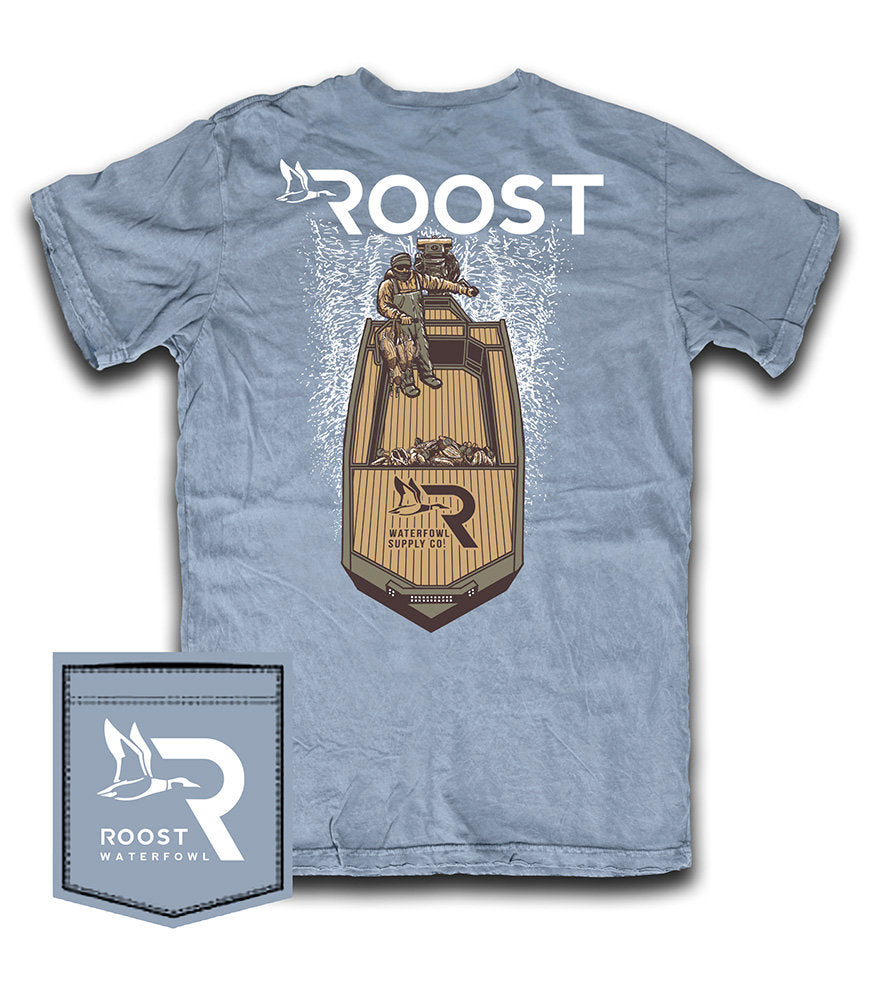 A light blue short-sleeve t-shirt with a large graphic on the back featuring a waterfowl and a wooden raft, with the word 'ROOST' printed above. A small matching fabric patch with the 'ROOST WATERFOWL' logo is shown in the bottom-left corner against a white background.