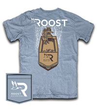 A light blue short-sleeve t-shirt with a large graphic on the back featuring a waterfowl and a wooden raft, with the word 'ROOST' printed above. A small matching fabric patch with the 'ROOST WATERFOWL' logo is shown in the bottom-left corner against a white background.