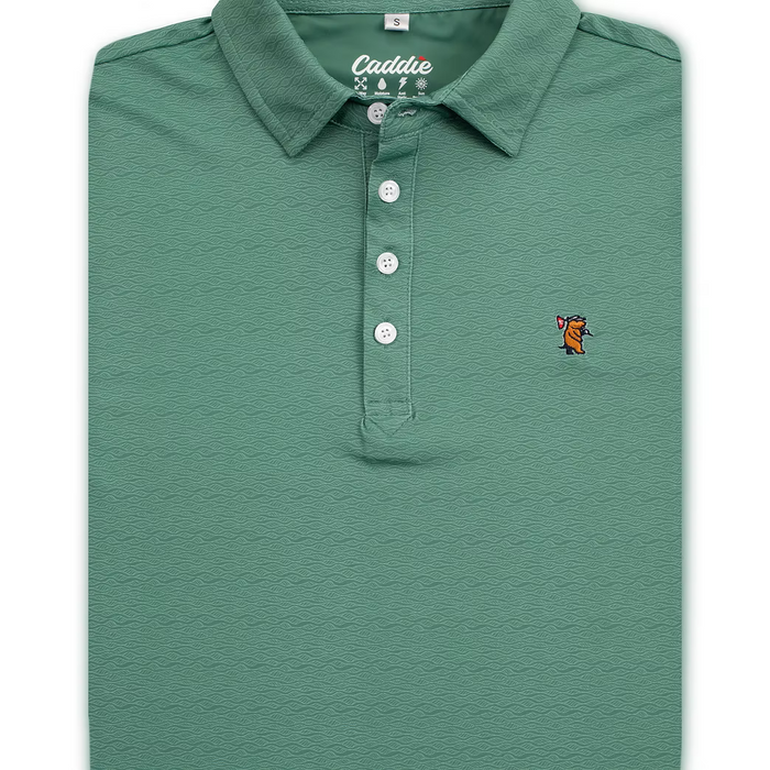 A folded sage green polo shirt with a textured wavy pattern, featuring a white button placket and a small embroidered fox logo on the left chest.