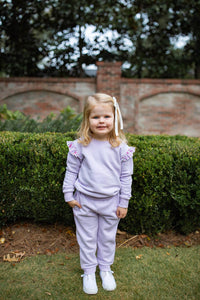 A young girl with blonde hair tied back with a white bow stands outdoors in front of a green hedge and brick wall, wearing a light lavender matching sweatshirt and jogger pants set with white sneakers.