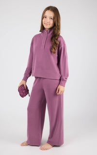 A young girl modeling a matching mauve purple lounge set consisting of a half-zip long-sleeve top and wide-leg pants, standing barefoot against a light gray studio background.