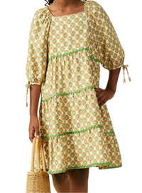 A Girl wearing a tiered short dress with a green and yellow geometric pattern on a beige background, holding a woven straw bag against a plain white background.