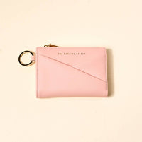 A light pink leather zip-around card holder with an envelope-style flap and a gold-tone key ring attachment, shown on a warm cream background. The brand name 'THE DARLING EFFECT' is printed in gold on the front.