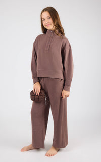 A young girl modeling a matching two-piece lounge set in a dusty mauve-brown color, consisting of a long-sleeve mock-neck top and wide-leg pants, standing against a light gray studio background.