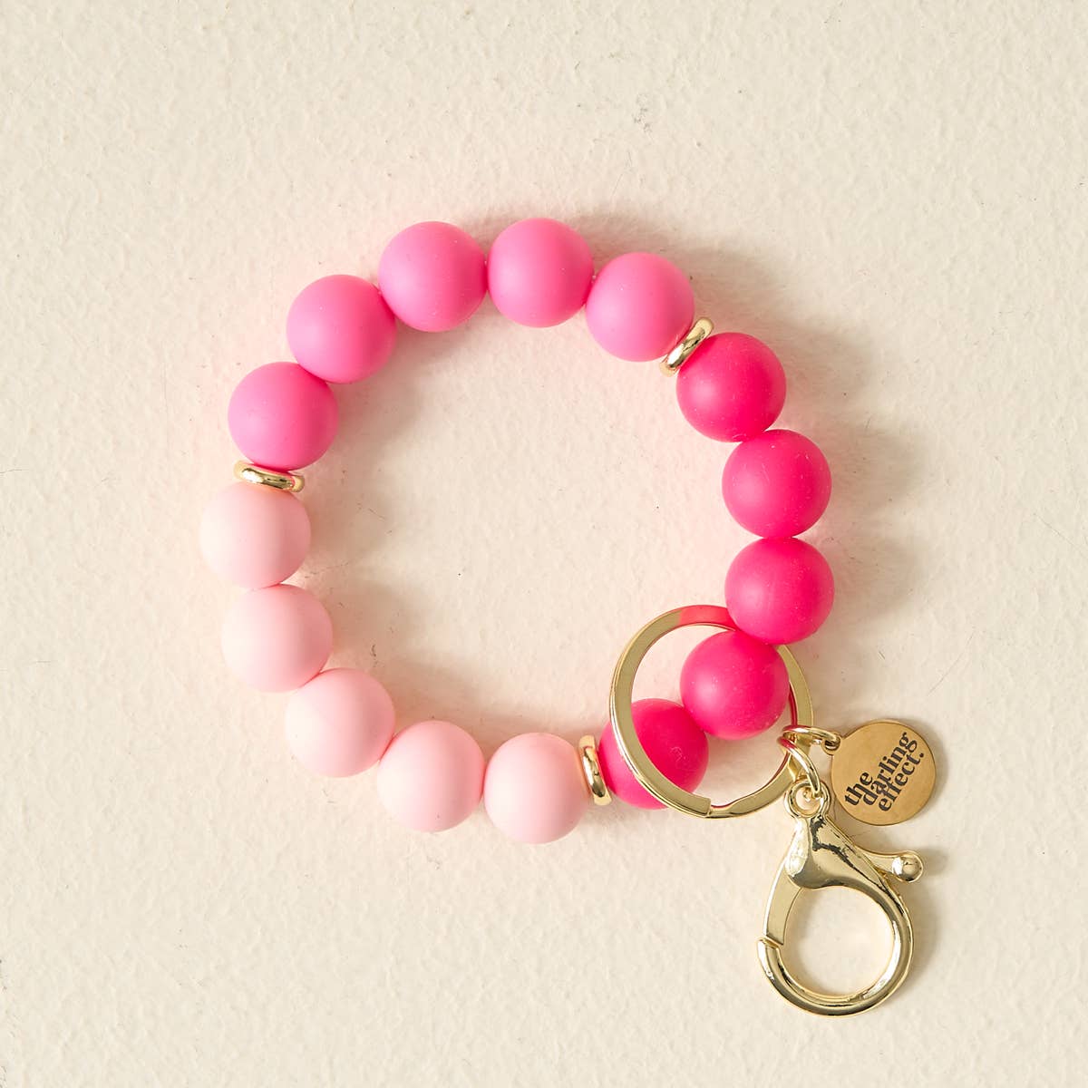 A beaded stretch bracelet in shades of pink, featuring a gold-tone split ring, lobster clasp, and a round charm engraved with 'One Garden, One Dream'.