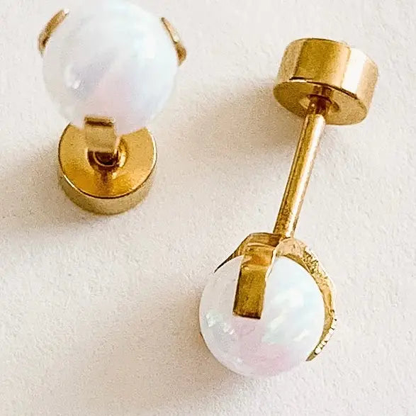 A pair of gold stud earrings with round, iridescent white pearl-like beads, shown on a light textured background.
