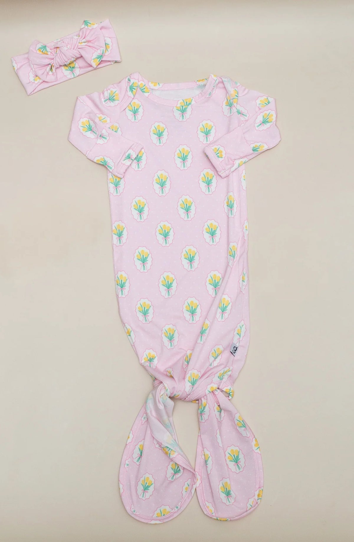 A baby footie sleeper in light pink with a repeating tulip print, shown with a matching pink bow headband on a light beige background.