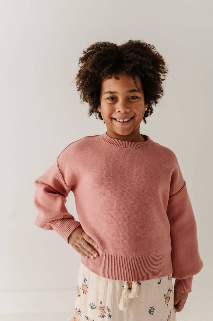 A smiling young girl with curly dark hair wearing a dusty rose pink knit sweater and a cream floral skirt, standing against a plain light gray studio background.