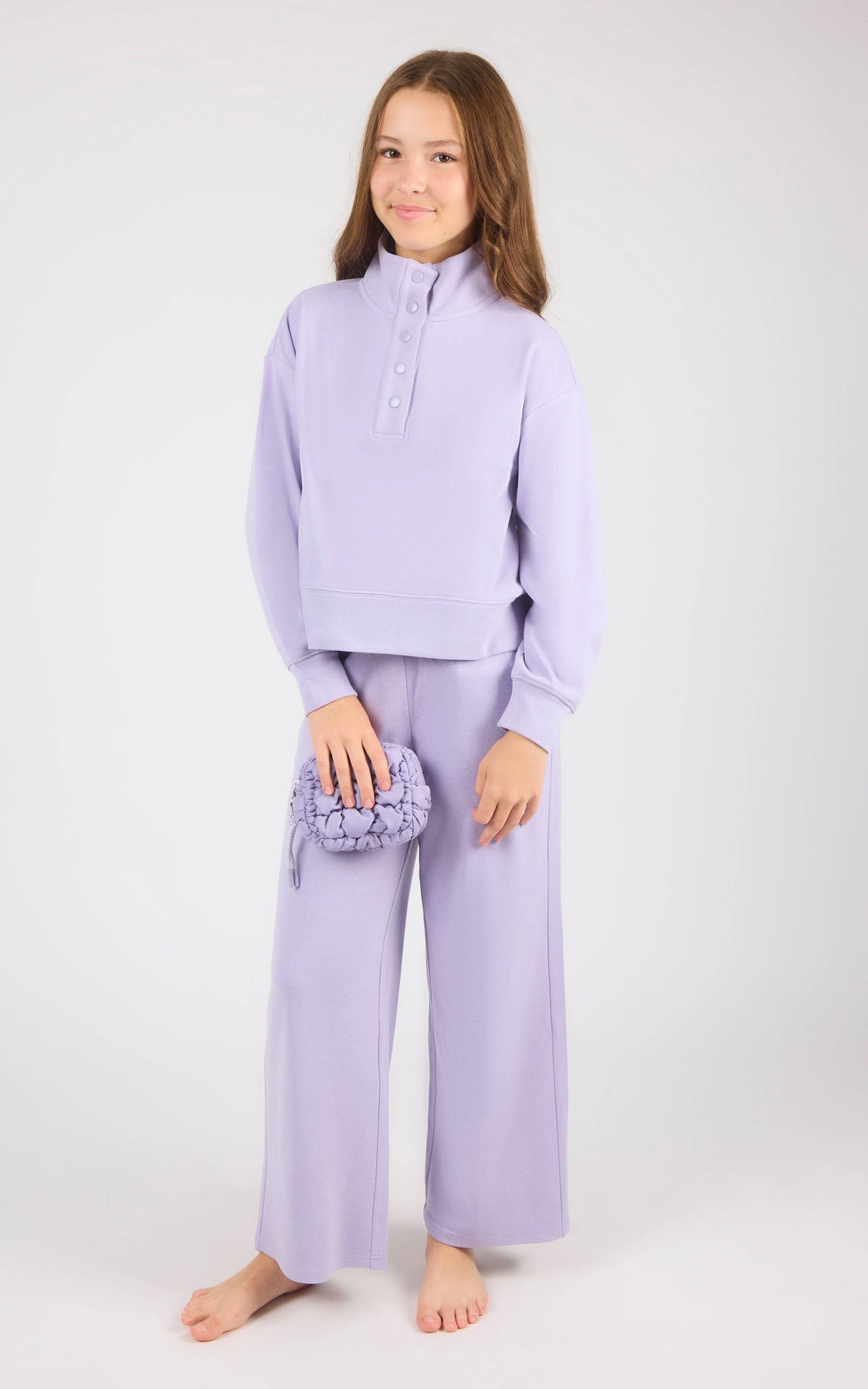 A young girl stands barefoot against a light gray studio background, wearing a matching lavender lounge set consisting of a cropped long-sleeve henley top and wide-leg pants, and holding a small textured lavender pouch.