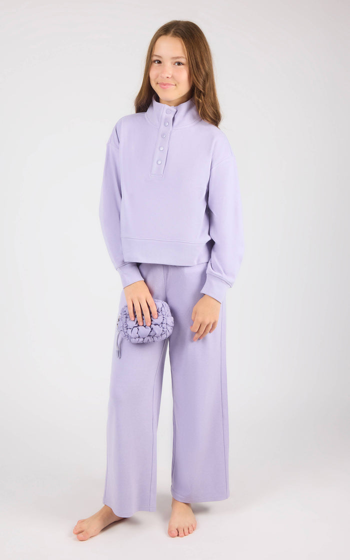 A young girl stands barefoot against a light gray studio background, wearing a matching lavender lounge set consisting of a cropped long-sleeve henley top and wide-leg pants, and holding a small textured lavender pouch.