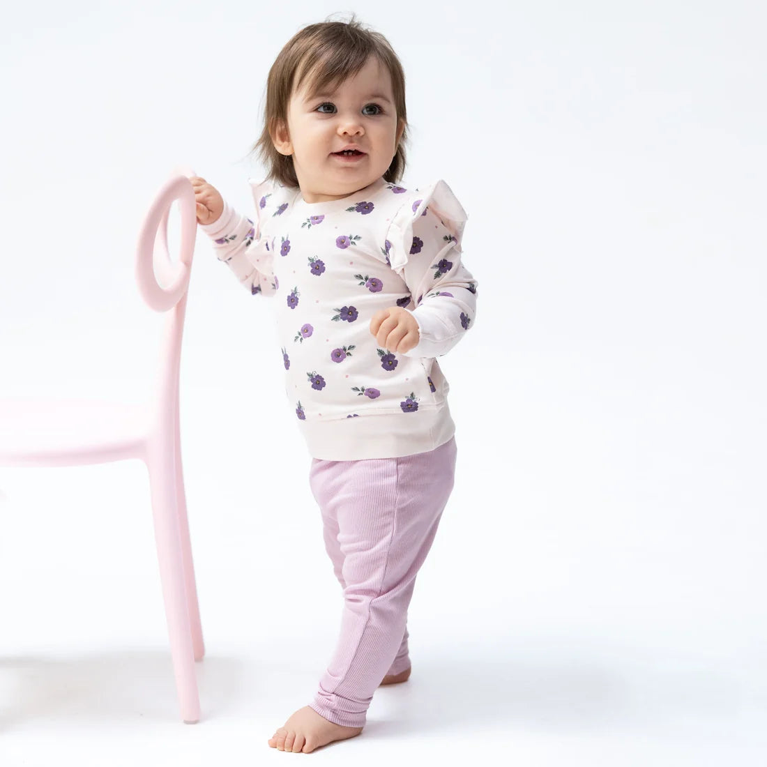 A toddler standing barefoot and holding a pink chair, wearing a cream long-sleeve top with a purple berry print and light pink pants, photographed against a clean white studio background.