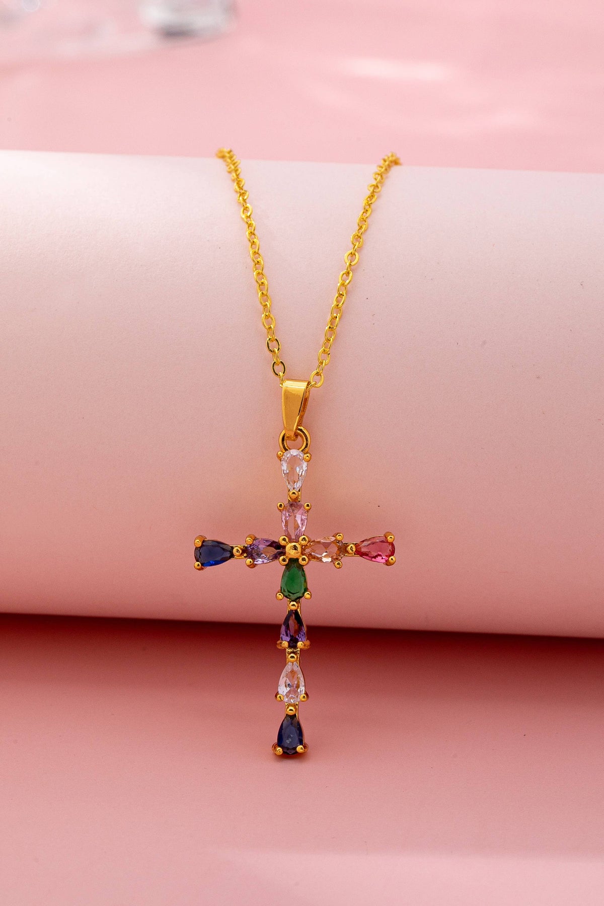 Gold cross necklace with colorful gemstones on a pink background