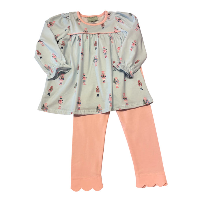 A toddler girl's two-piece outfit featuring a long-sleeve smocked blouse in sage green with a nutcracker print, paired with peach leggings with scalloped hems.