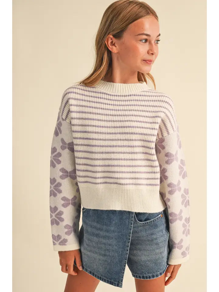 A young girl wearing a cream knit sweater with purple horizontal stripes on the chest and purple floral patterns on the sleeves, paired with denim shorts, set against a solid beige background.
