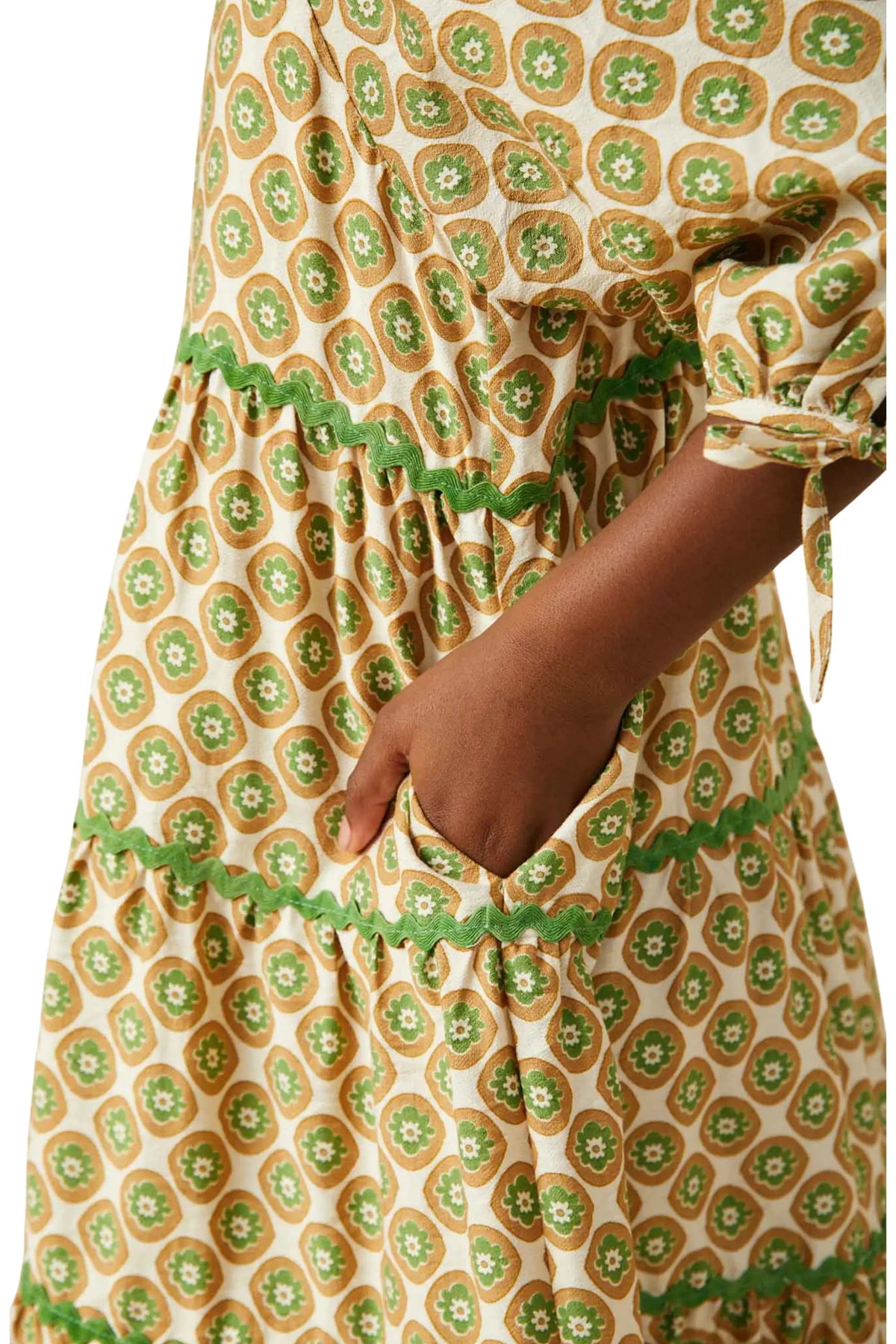Close-up of a tiered dress with a green and brown circular floral pattern on a cream background, featuring a hand in a side pocket and green lace trim.