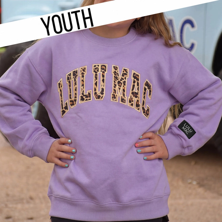 A youth model wearing a lavender purple crewneck sweatshirt with 'LULUMAC' printed in leopard print across the chest, featuring a black sleeve patch with 'LULU MAC' text. A white diagonal banner with 'YOUTH' is in the top left corner.