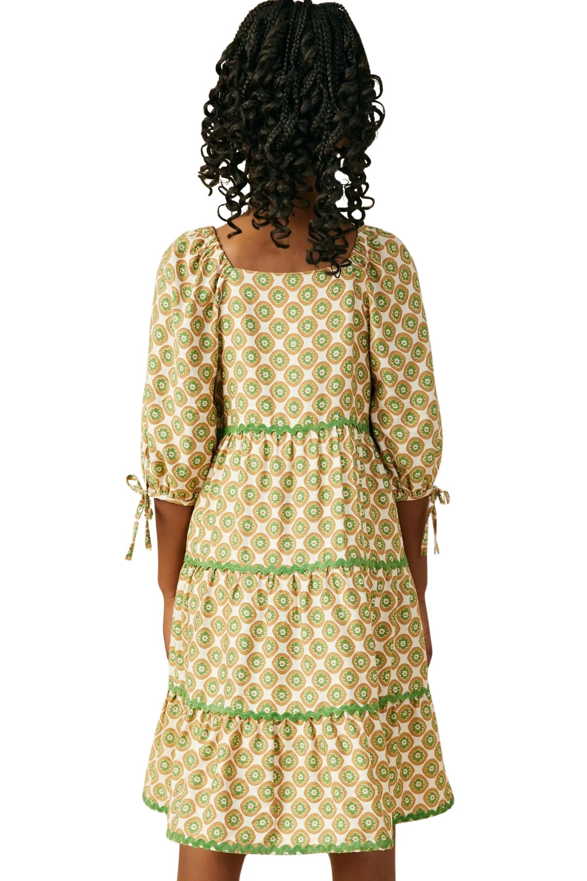 A woman shown from the back wearing a tiered midi dress with a green and beige geometric pattern, 3/4 puff sleeves, and tie details at the cuffs, set against a plain white background.