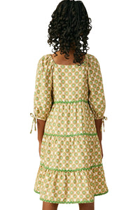 A woman shown from the back wearing a tiered midi dress with a green and beige geometric pattern, 3/4 puff sleeves, and tie details at the cuffs, set against a plain white background.